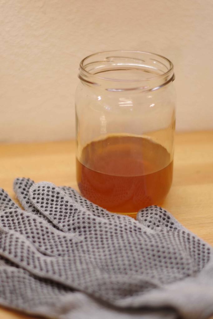 Ein Glas, in dem Bienenwachs im Ofen geschmolzen wurde. Davor liegen zwei Handschuhe, um die Finger vor Verbrennung zu schützen.